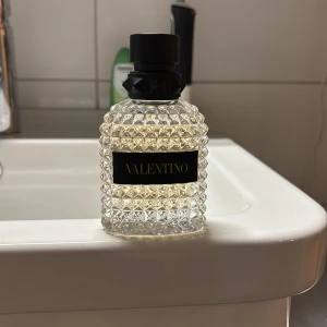 Valentino parfym - Elegant parfym från Valentino (med box) i en stilren, glasklar flaska med ett mönster av små pyramider. Flaskan har en svart etikett med guldtext och en matchande svart kork. Perfekt för den som vill ha en lyxig doftupplevelse.