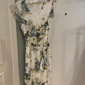 Blommig klänning med volangdetaljer - Charmig blommig klänning i ljusa toner med volangdetaljer och knytband i midjan. Perfekt för sommarens festligheter.