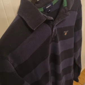 Randig pikétröja från GANT - Säljer en snygg randig pikétröja från GANT i mörkblått och svart med gröna detaljer på kragen. Tröjan har långa ärmar och en klassisk pikékrage. Perfekt för en stilren look!