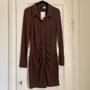 Brun klänning med knappar - Helt ny prislapp kvar. Snygg skjort liknande brun klänning. Den har en stilren design med rynkade detaljer längs knappraden som ger en fin passform. Perfekt för en chic och elegant look.