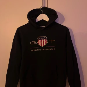 GANT hoodie  - Säljer en svart hoodie från GANT med deras klassiska logga och texten 'American Sportswear' på framsidan. Hoodien har en bekväm passform och är perfekt för en avslappnad stil. Den är tillverkad i ett mjukt material som håller dig varm och skön.