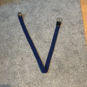 Blått flätat bälte - Snyggt blått flätat bälte med silverfärgat spänne. Perfekt för att ge en stilren touch till din outfit. Bältet är flexibelt och passar olika storlekar.