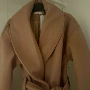 Beige kappa med bälte - Säljer en elegant beige kappa med bälte. Den har en klassisk design med bred krage och långa ärmar. Perfekt för att hålla stilen under kyligare dagar.