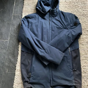 Mörkblå jacka från Hugo Boss - Säljer en stilren mörkblå jacka från Hugo Boss. Jackan har en praktisk huva och dragkedja framtill. Den är vattenavvisande och lätt, perfekt för varierande väder. Designad med långa ärmar och diskreta fickor med dragkedjor.