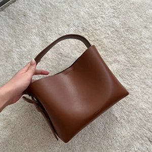 ARKET- Brun handväska i skinn - Snygg brun handväska i skinn med ett stilrent och minimalistiskt utseende. Väskan har ett praktiskt handtag och en justerbar axelrem. Den har en elegant bältesdetalj på sidan. ❤️ Den är i jätte fint skick utan defekter!