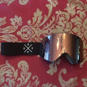 Skidglasögon från Dope - Säljer mina snygga svarta skidglasögon från Dope med spegelglas i rosa färg. De har en bred, justerbar rem med logotyp. Perfekta för vinterns äventyr i backen. Kommer med ett praktiskt fodral för säker förvaring. Inga repor eller skador på glaset. Endast ett litet slitage på bandet i form av lite nopprigt (syns på bilden). Nypris 1299kr.  Skriv vid frågor 🤩