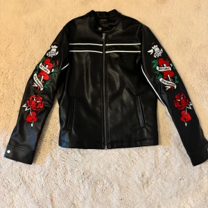 Svart skinnjacka från Ed Hardy - Snygg svart skinnjacka från Ed Hardy med coola broderade detaljer i rött och vitt. Jackan har långa ärmar och dragkedja framtill. Perfekt för dig som vill sticka ut med stil! Pris går att diskutera :)