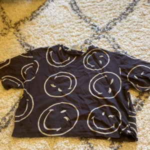 Svart t-shirt med smiley-mönster från H&M - Svart t-shirt från H&M med stora vita smiley-ansikten över hela tyget. Perfekt för en avslappnad stil. Storlek 158/164💗