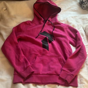 Rosa hoodie från Peak Performance - Säljer en snygg rosa hoodie från Peak Performance med svart logga på framsidan. Den har en justerbar huva med svarta snören och en praktisk magficka. Perfekt för en avslappnad stil!
