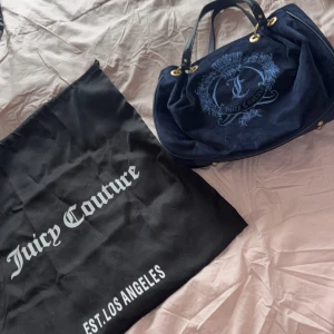 Mörkblå axelväska från Juicy Couture - Snygg mörkblå axelväska från Juicy Couture med broderad logga och gulddetaljer. Väskan har ett rymligt fack med mönstrat innerfoder och dragkedja. Perfekt för att ge din outfit en lyxig touch.