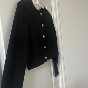 Svart kavaj med knappar - Snygg svart kavaj med silverfärgade knappar framtill. Den har en klassisk design med långa ärmar och två fickor. Perfekt för en stilren look.