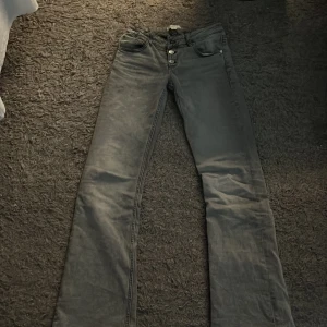 Grå jeans från Gina Tricot - Snygga grå jeans från Gina Tricot med bootcut-stil och hög midja. Byxorna har en klassisk femficksdesign och stängs med tre knappar framtill. Perfekta för en avslappnad look.