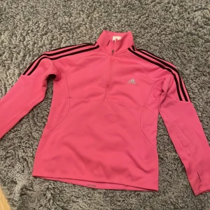 Rosa träningsjacka från Adidas - Säljer en snygg rosa träningsjacka från Adidas med svarta ränder på ärmarna. Jackan har en halv dragkedja framtill och är perfekt för träning eller vardagsbruk. Den är i mycket bra skick och har en bekväm passform. Perfekt för höst och vår!