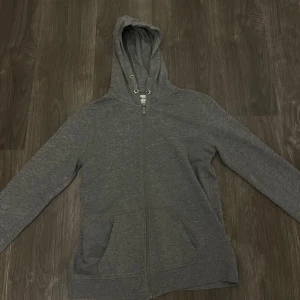 Grå hoodie med dragkedja - Säljer en grå hoodie i mjukt material med dragkedja framtill. Perfekt för vardagsbruk eller mysiga dagar hemma. Den har en klassisk passform och en praktisk huva. Fickor på sidorna för extra bekvämlighet. 💞💕 