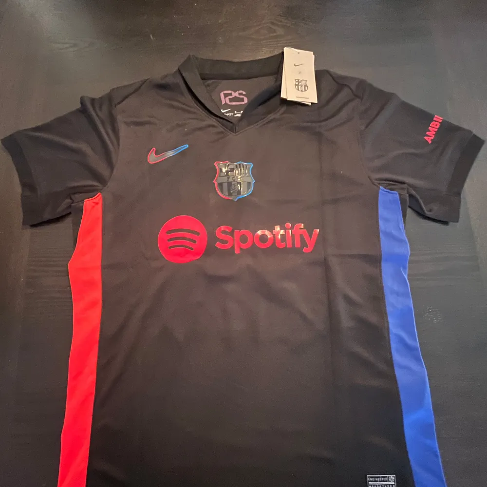 Säljer en svart FC Barcelona fotbollströja i nytt skick. Den har korta ärmar och coola röda och blå detaljer på sidorna. Framtill finns Spotify-loggan och Nike-märket. På baksidan står det 'Lamine Yamal' och nummer 19 i rött. Perfekt för alla Barca-fans! 💙❤️. T-shirts.