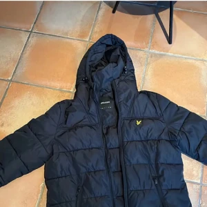Marinblå  pufferjacka från Lyle & Scott - Säljer en marinblå pufferjacka från Lyle & Scott i mycket bra skick. Jackan har en praktisk huva och dragkedja framtill. Perfekt för kyliga dagar och håller dig varm och stilren. Passar både till vardags och lite finare tillfällen. 🖤
