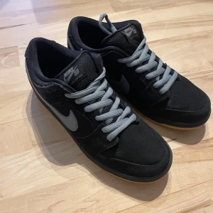 Nike SB Dunk Low Pro i svart och grå - Säljer ett par Nike SB Dunk Low Pro i svart och grått. Skorna har en snygg design med grå snörning och en gummisula i gult. Perfekta för både vardag och skate. De är i mycket bra skick och kommer i originalförpackning. Skriv om ni har frågor eller funderingar! Pris ej hugget i sten!!