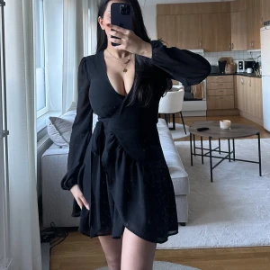 Svart klänning med v-ringning - Snygg svart klänning med djup v-ringning och långa ärmar. Den har en volangdetalj i kjolen som ger en fin rörelse. Perfekt för festliga tillfällen! Klänningen är i mycket bra skick och passar bra till både höst och vår.