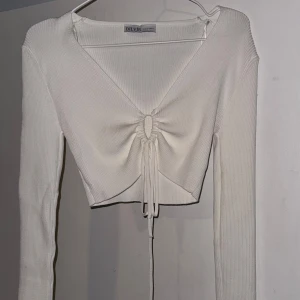 Vit ribbad topp från Dilvin - Säljer en superfin vit ribbad topp från Dilvin. Den har en snygg v-ringning och knytning framtill som ger en cool look. Toppen är croppad och har långa ärmar, perfekt för både vardag och fest. Passar bra till jeans eller kjol! Storlek S