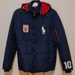 Sommar25. Dunjacka från Polo Ralph Lauren L. - Säljer en snygg blå dunjacka från Polo Ralph Lauren. Den har en broderad logga på bröstet och nummer 10 på ärmen. Jackan är långärmad med dragkedja och har en röd insida i huvan. Den har mest hängt i garderoben och blivit avglömd stl L.
