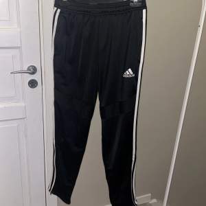 Säljer ett par svarta träningsbyxor från Adidas i storlek 164. Byxorna har vita ränder längs sidorna och en elastisk midja för bekväm passform. Perfekta för träning eller vardagsbruk. Adidas-loggan är tryckt på framsidan. De är i mycket bra skick och redo för nya äventyr!