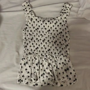 Vit topp med palmmönster från H&M - Säljer en vit topp från H&M med svart palmmönster. Toppen har en smockad överdel och volangdetalj nedtill. 