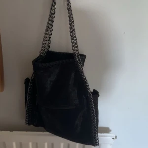 Svart axelväska med kedjedetaljer - Säljer en snygg svart axelväska i mycket bra skick. Väskan har coola kedjedetaljer och en praktisk dragkedja. Perfekt för både vardag och fest!