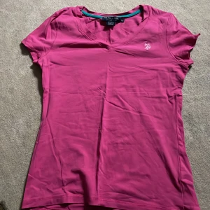 Rosa t-shirt från U.S. Polo Assn. - Säljer en snygg rosa t-shirt från U.S. Polo Assn. i storlek XS. Den är gjord av bomull och spandex för en skön och stretchig passform. T-shirten har en v-ringning och ett broderat logotypmärke på bröstet. Köpt i New York
