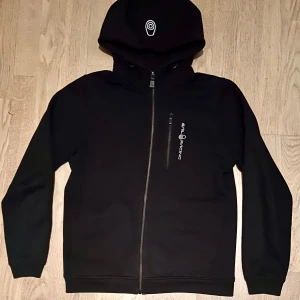 Svart hoodie från Sail Racing - Säljer en svart hoodie från Sail Racing i mycket bra skick. Den har en dragkedja framtill och en justerbar huva. Perfekt för höst och vår, med en stilren design och bekvämt material. Passar både till vardags och lättare aktiviteter.