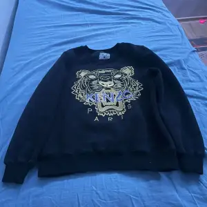 Säljer en svart sweatshirt från Kenzo med ett coolt tigerbroderi i guld och blått på framsidan. Tröjan har långa ärmar och är i mycket bra skick. Perfekt för höst och vinter, och den är superbekväm att ha på sig. Passar både till vardags och fest!