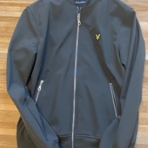 Grön bomberjacka från Lyle & Scott - Säljer en snygg grön bomberjacka från Lyle & Scott i mycket bra skick. Jackan har en dragkedja framtill och två fickor med dragkedjor. Den är perfekt för höst och vår, med ett stilrent broderat emblem på bröstet. Passar både till vardags och fest!