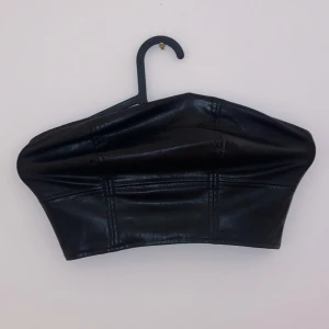 Svart tubtopp i skinnimitation - Snygg svart tubtopp i skinnimitation som ger en cool och edgy look. Perfekt för en utekväll eller fest. Toppen är tight och sitter bra på plats. Passar perfekt till både jeans och kjol för en stilren outfit.