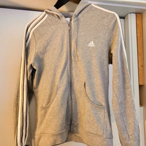 Grå hoodie från Adidas - Säljer en grå hoodie från Adidas i mycket bra skick. Den har de klassiska tre ränderna längs ärmarna och en dragkedja framtill. Perfekt för både träning och vardag. Superbekväm och stilren design!