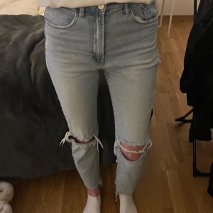 Ljusa jeans med slitningar - Säljer ett par ljusa högmidjade mom slim fit jeans från Stradivarius med snygga slitningar på knäna. Det enda negativa är att det fanns ett hål på ena höften redan som nya, men har sytt ihop det. Använd fåtal gånger