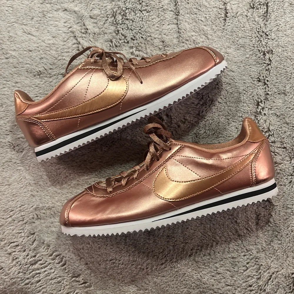 Snygga Nike Cortez sneakers i en glansig roséguld färg. De har en klassisk design med snörning och en vit sula med svart rand. Perfekta för att ge din outfit en extra touch av stil. Skorna är i ny skick och redo för nya äventyr. Storlek 38,5 EU för kvinna. Skor.