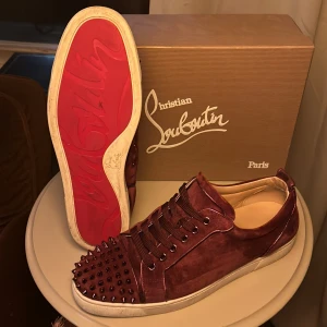 Christian Louboutin - Säljer ett par snygga vinröda sneakers från Christian Louboutin i mocka. Skorna har den ikoniska röda sulan och är dekorerade med coola nitar på tån. Perfekta för att få många blickar! De är i mycket bra skick och inga spikar saknas😇❤️                                                                                      OBS! Endast skorna hänger med