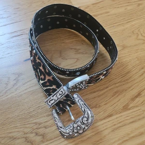 Leopardmönstrat bb belt från Zara - Säljer ett coolt leopardmönstrat bälte från Zara med en stort, detaljerat silverspänne. Bältet har glittriga stenar och nitar längs kanterna som ger en extra touch av glamour. Perfekt för att piffa upp vilken outfit som helst! 🐆✨
