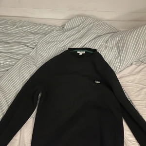 Svart hoodie från Lacoste - Säljer en svart Lacoste hoodie i M. Skicket är som nytt. Har loggan på framsidan och även ett snyggt mönster på bägge sidorna av armarna 
