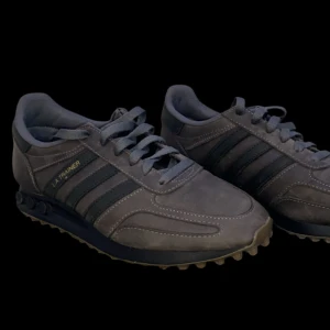 Grå Adidas LA Trainer sneakers - Säljer ett par grå Adidas LA Trainer sneakers i snygg retrostil. Skorna har de klassiska tre ränderna i svart på sidan och är gjorda i mocka. Perfekta för både vardag och träning. De har en bekväm passform och snygg design som funkar året runt. Skorna har små tecken för använding (en lite större på ena skons framsida). Vid frågor eller funderingar så är det bara att skriva😃👍