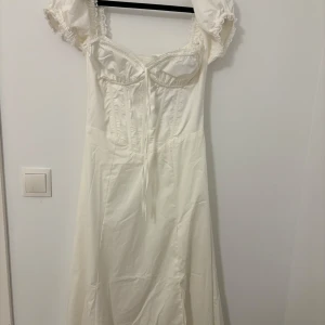 Vit klänning med puffärmar - Charmig vit klänning med puffiga kortärmar och en söt knytning framtill. Klänningen har en romantisk stil med spetsdetaljer vid ärmslut och en smickrande passform. Perfekt för sommarens fester eller en midsommarnattsdröm. Materialet känns lätt och luftigt, vilket gör den idealisk för varma dagar.