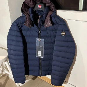 Colmar jacka (NY) - Säljer en snygg och varm mörkblå dunjacka från Colmar. Jackan har en praktisk dragkedja och en avtagbar huva med knappar. Perfekt för kalla vinterdagar. Den är i nytt skick med etikett kvar. Passar både till vardags och mer uppklädda tillfällen.