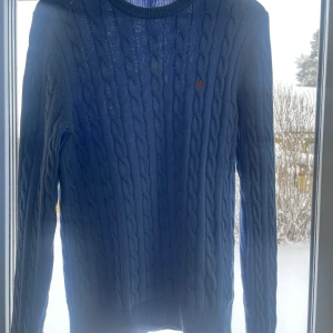 Blå kabelstickad tröja från Morris - Säljer en snygg blå kabelstickad tröja från Morris i storlek S. Den är gjord av 100% Pima bomull vilket gör den supermjuk och bekväm. Perfekt för både höst och vår! Tröjan har långa ärmar och en klassisk rund halsringning. Den lilla loggan på bröstet ger en extra touch av stil. Tyvärr så har den en liten fläck som man ser i mitten!