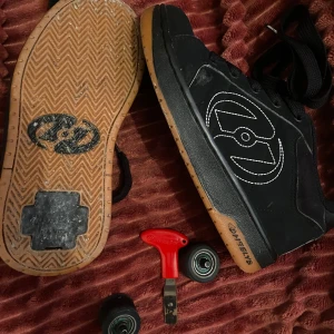 Svarta Heelys med hjul och verktyg - Säljer ett par svarta Heelys i bra skick. Skorna har en cool design med vita sömmar och kommer med hjul och verktyg för att enkelt byta. Perfekta för att rulla runt. Sulan är brun och slitstark.