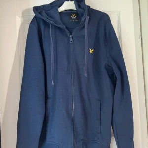 Blå hoodie från Lyle & Scott - Säljer en snygg blå hoodie från Lyle & Scott i bra skick. Den har en dragkedja framtill och en gul broderad logga på bröstet. Perfekt för både höst och vår, med en skön passform och praktiska fickor. Perfekt för vardagsbruk eller en avslappnad dag ute.