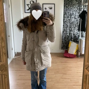 Beige parkajacka med pälsdetaljer - Säljer en snygg beige parajumperjacka med stor pälskantad huva. Perfekt för kalla vinterdagar! Jackan har flera praktiska fickor och stängs med både dragkedja och knappar. Den är i bra skick och supermysig att ha på sig. Passar perfekt för vinterns alla äventyr! ❄️