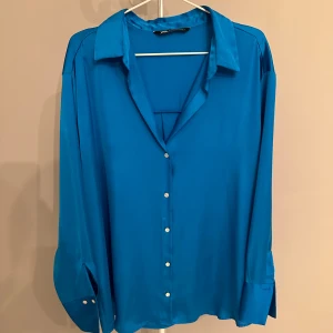 Blå långärmad skjorta från Zara - Säljer en snygg blå långärmad blus från Zara i storlek XXL. Skjortan har en klassisk krage och knappar framtill. Perfekt för både vardag och festliga tillfällen. Materialet känns mjukt och bekvämt. Passar bra till både jeans och kostymbyxor! I material som påminner om silke 