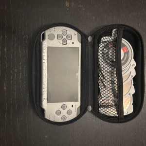 Sony PSP med spel och fodral - Saljer en silverfargad Sony PSP med ett gäng spel. Konsolen kommer i ett praktiskt svart fodral med dragkedja. Perfekt för dig som älskar retro gaming! Spelen inkluderar titlar som Grand Theft Auto och Tekken. Allt är i bra skick och redo för nya äventyr. Batteri ingår ej 