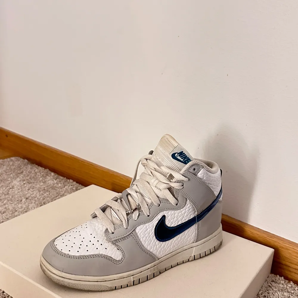 Säljer ett par Nike Dunk sneakers i vitt och grått med en snygg blå swoosh. Skorna har en klassisk design med snörning och är i mycket bra skick. Perfekta för både vardag och festliga tillfällen!. Skor.