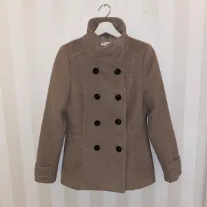 Säljer en snygg beige kappa från H&M i storlek 38. Den har en dubbelknäppt design med svarta knappar och en hög krage. Perfekt för höst och vår! 🧥