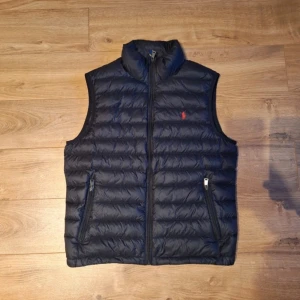 Mörkblå dunväst från Polo Ralph Lauren - Säljer en snygg svart dunväst från Polo Ralph Lauren. Den är perfekt för höst och vår, och har den klassiska röda Polo-loggan på bröstet. Väldigt stilren och lätt att matcha med andra plagg!